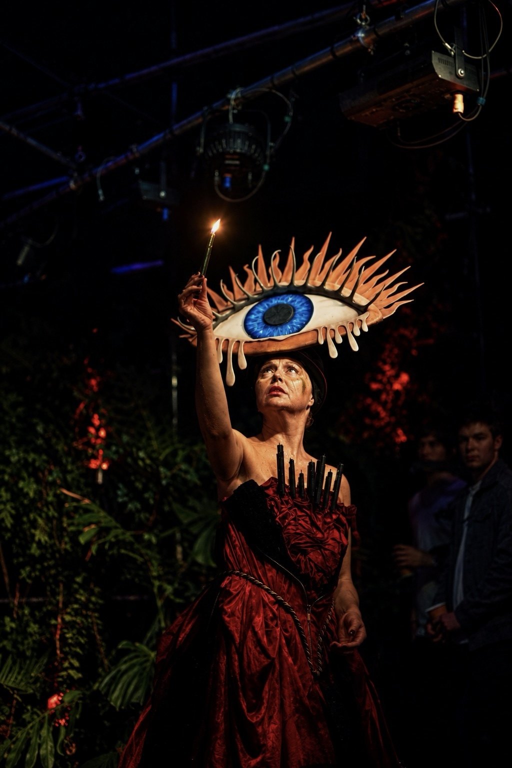 Laura King holding a lit candle above her head, in the eye headdress and a red gown.
