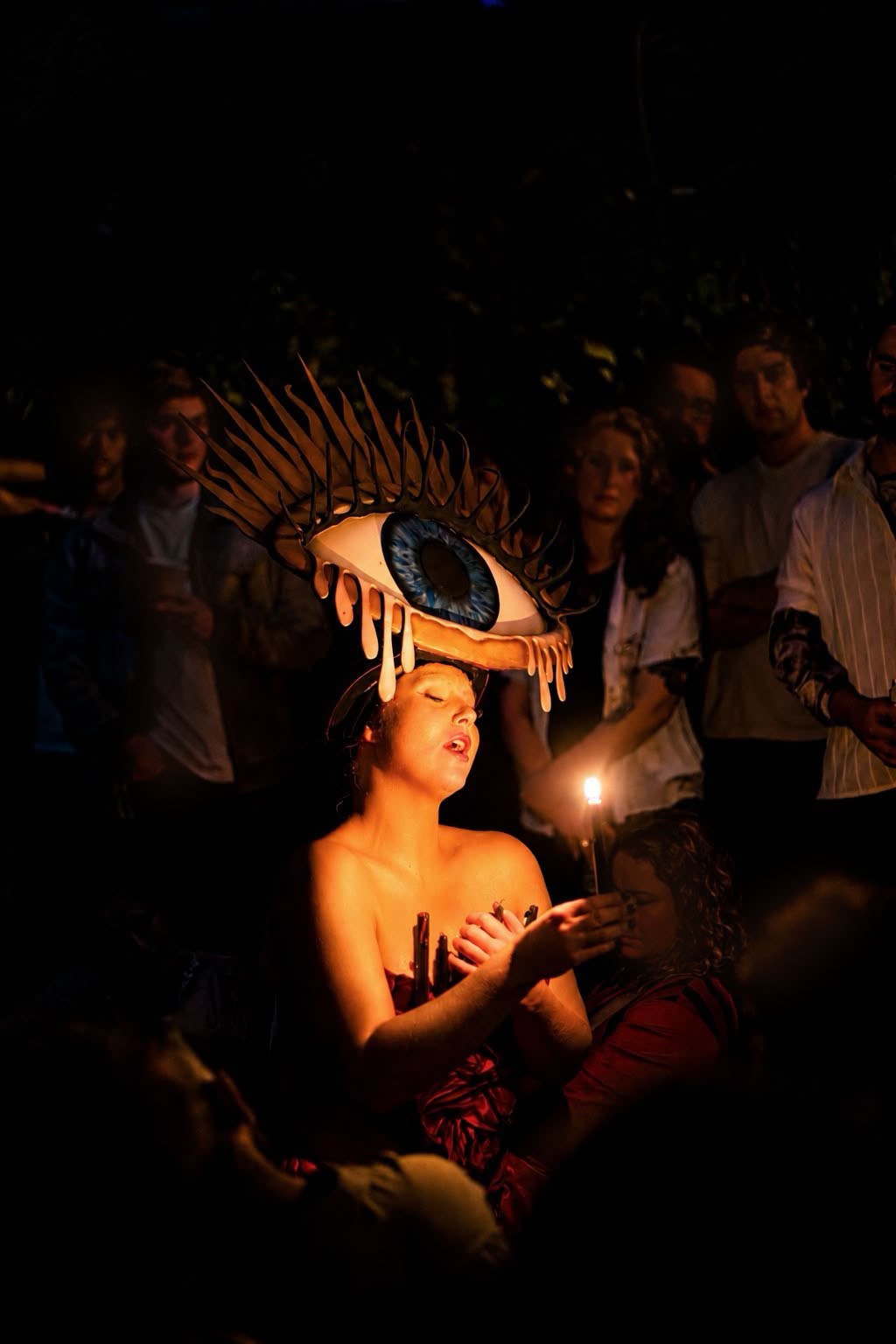 Laura King performing at night by candlelight, wearing a large sculpted eye headdress with dripping tears, surrounded by an audience.